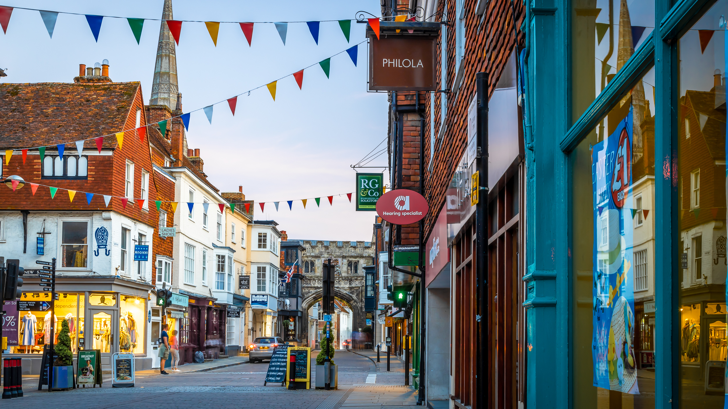 A charming UK street lined with shops and colorful bunting, featuring a historic archway in the background at dusk.