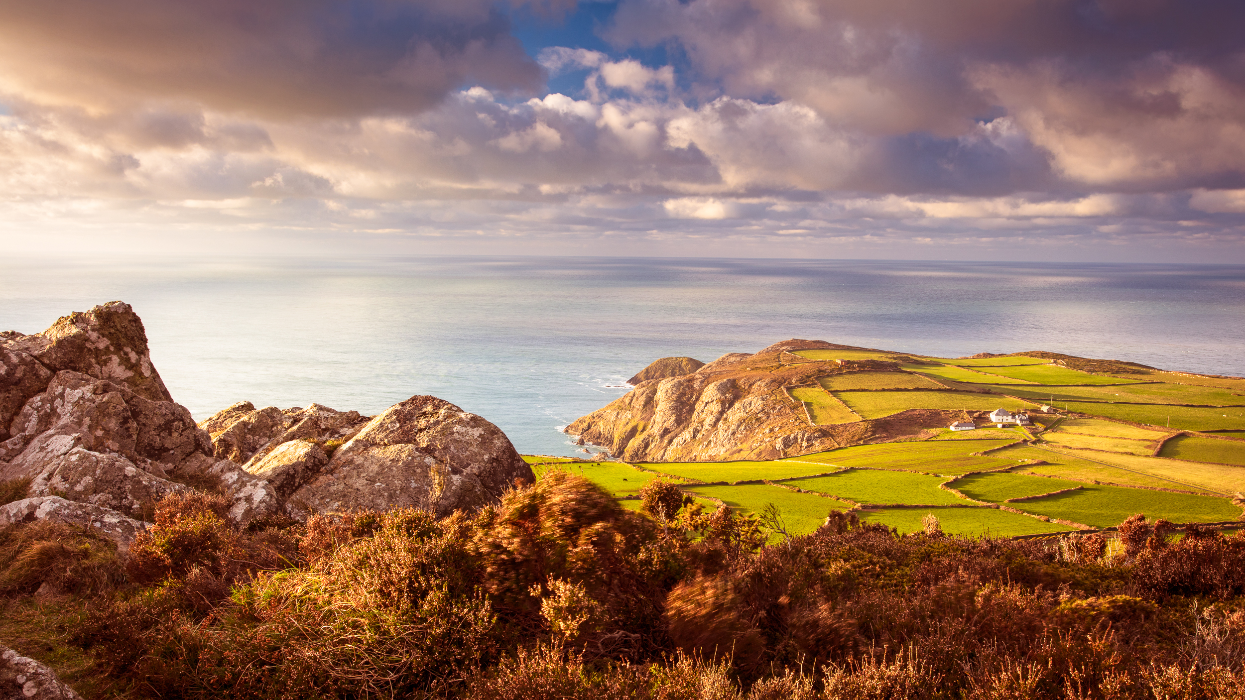 Stunning coastal landscape featuring rugged rocks, rolling green fields, and a tranquil sea under a colorful sky.