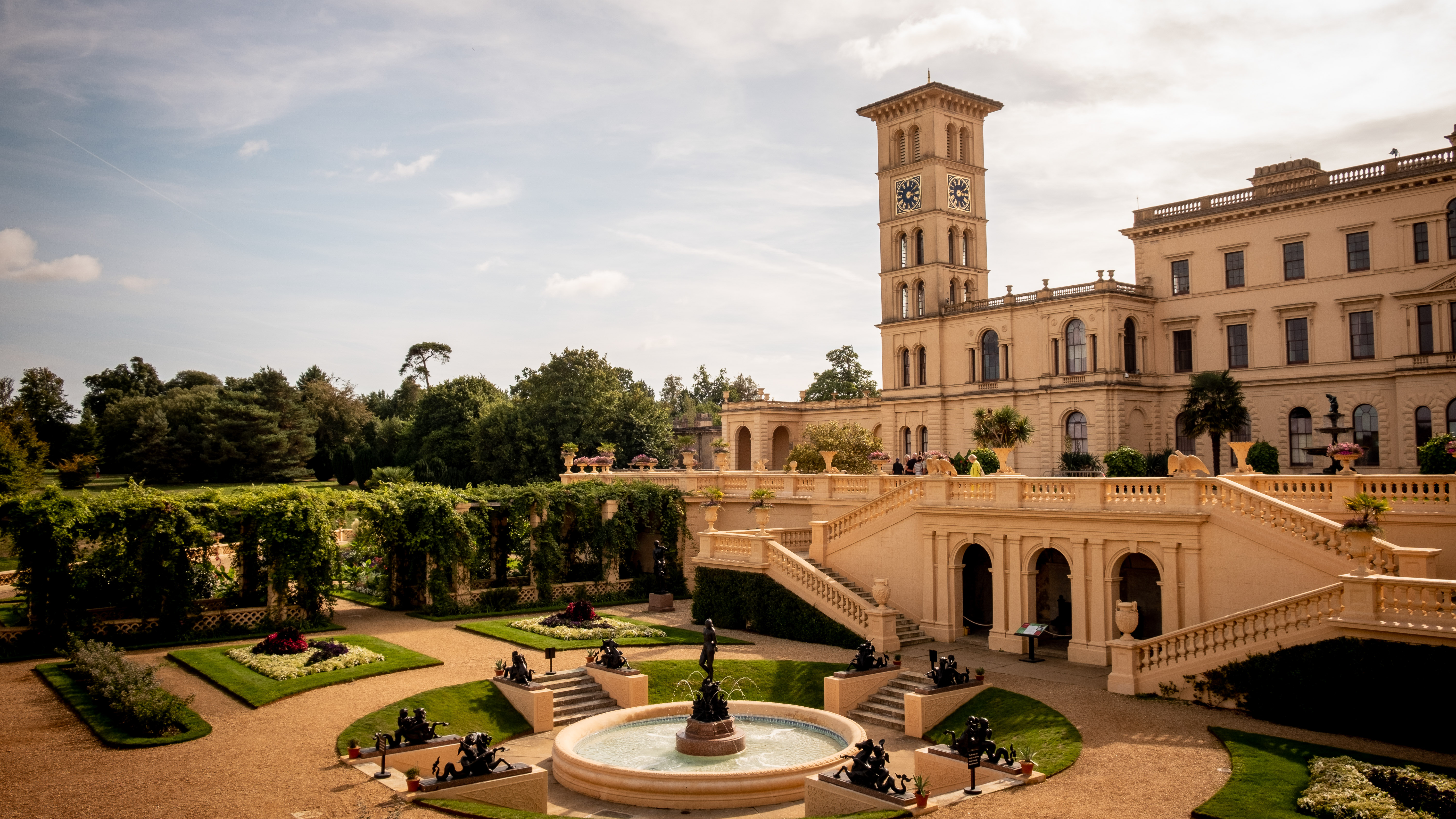 A grand building with a clock tower overlooks beautifully landscaped gardens featuring sculptures and fountains.