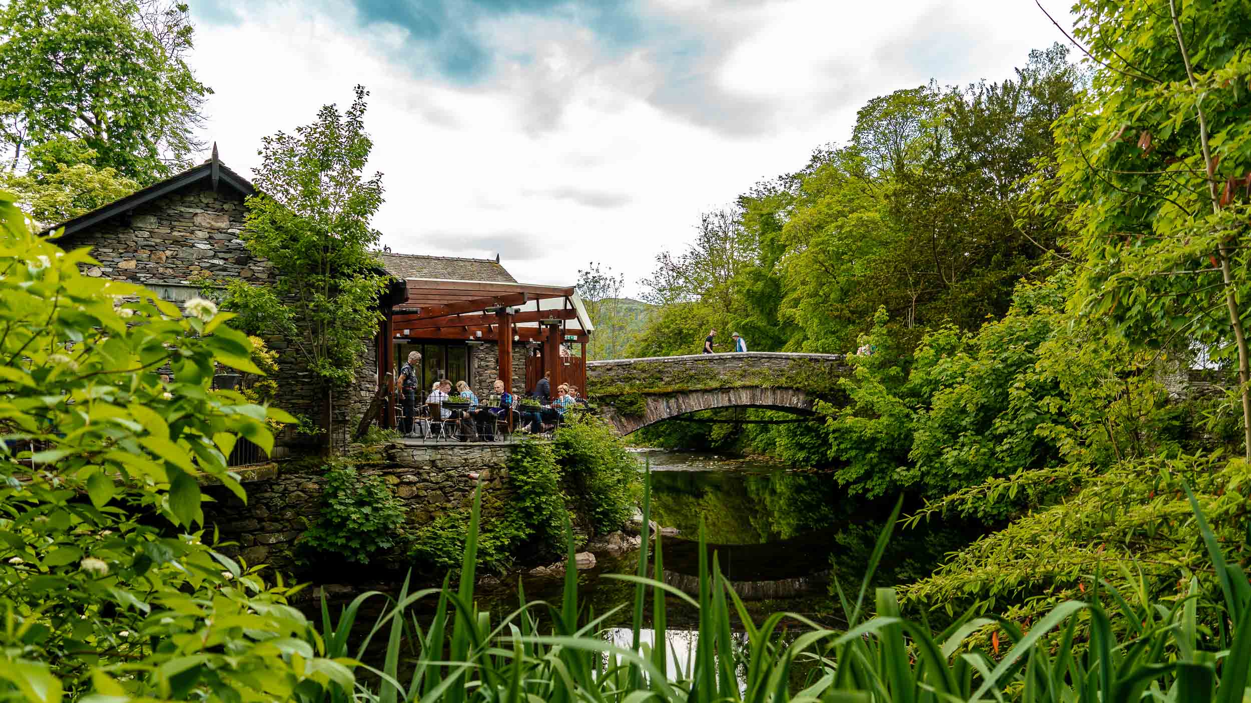 A scenic view of a quaint stone building with outdoor seating beside a tranquil stream and an arched stone bridge, surrounded by greenery.