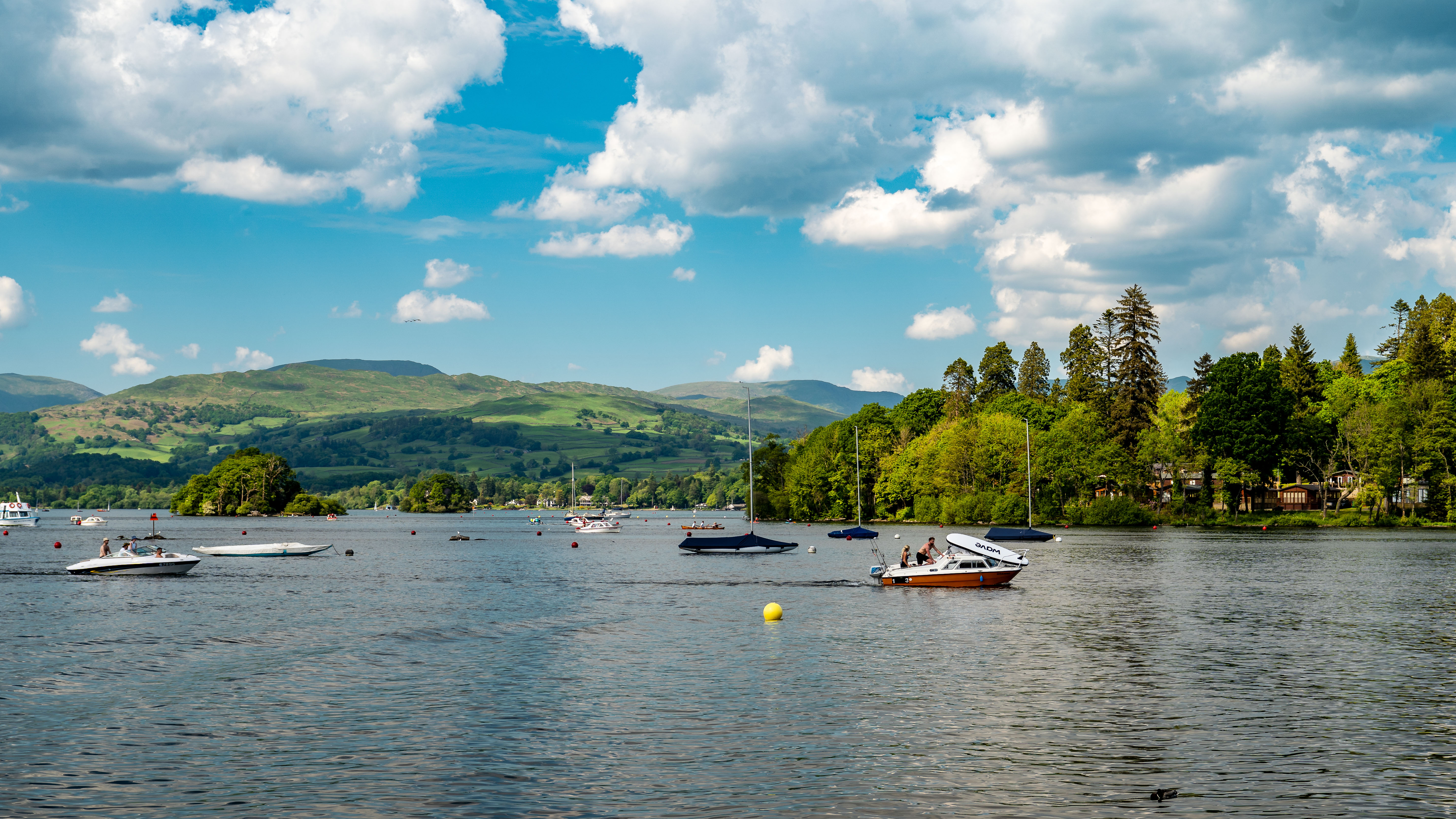 Scenic view of a lake with boats, surrounded by lush greenery and hills under a bright, partly cloudy sky.