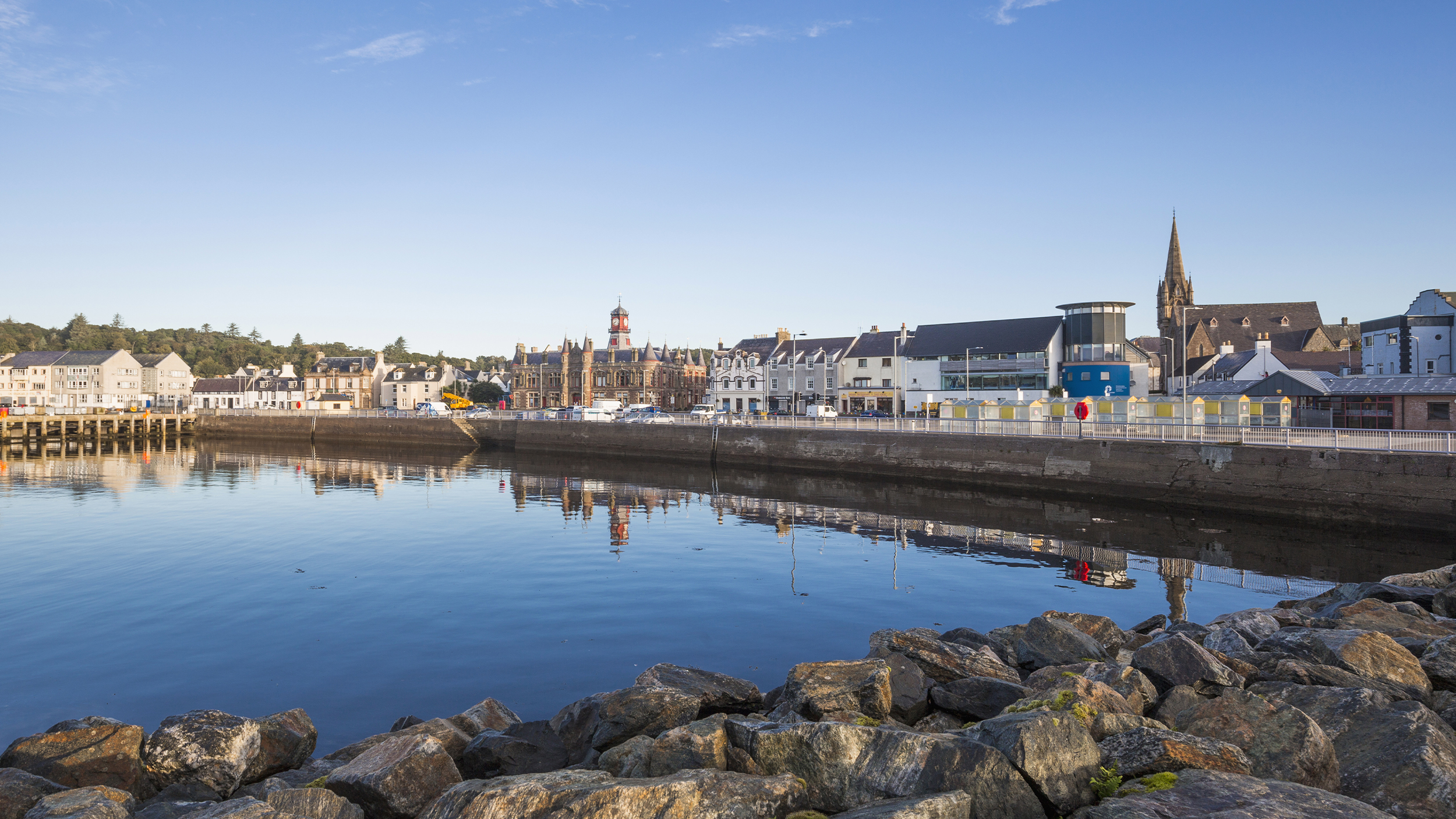 A tranquil waterfront scene showcasing buildings along the shore, reflecting in calm waters under a clear blue sky.