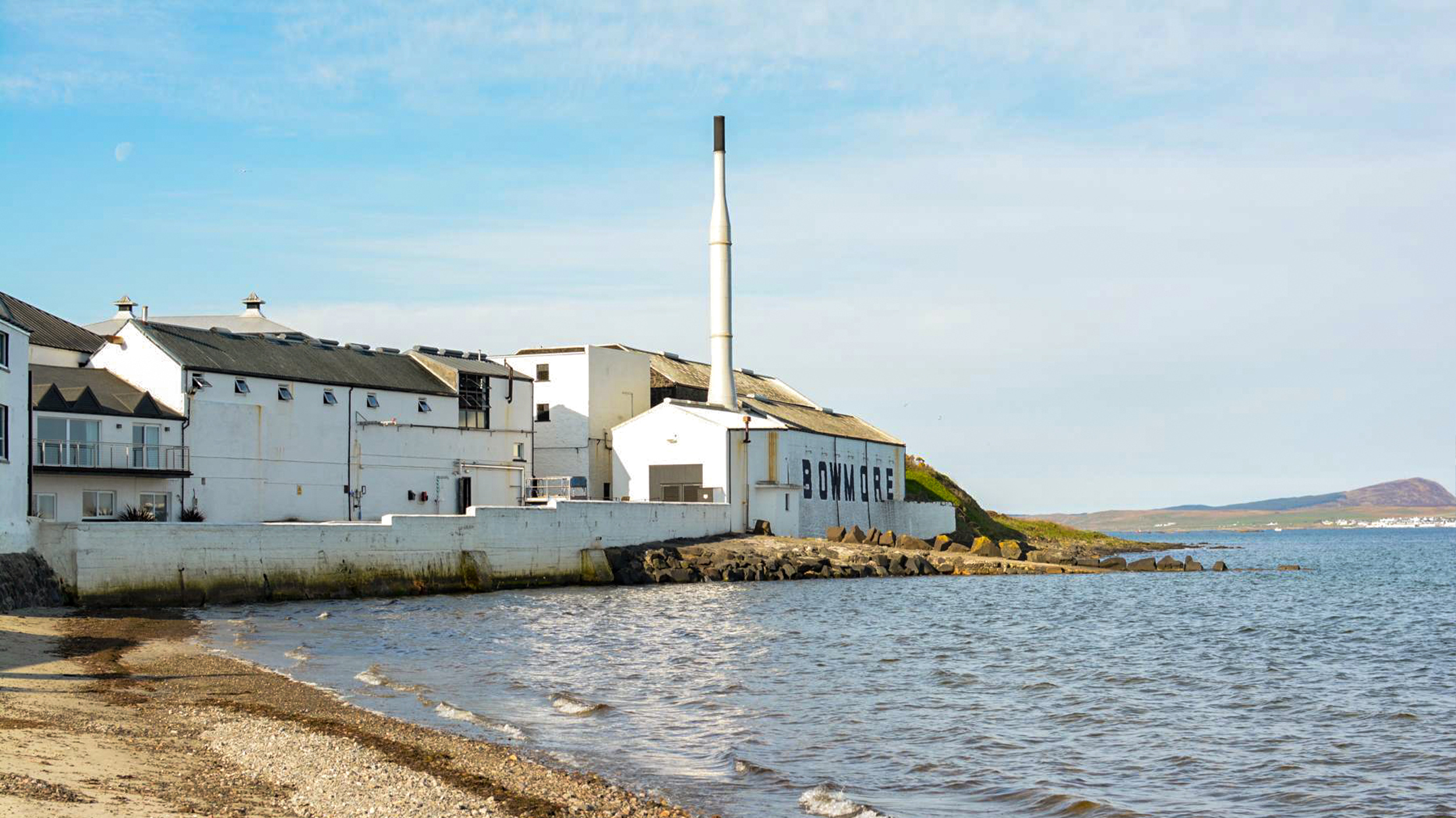 Coastal view of a distillery in Bowmore, with a white building and smokestack alongside gentle waves and a clear blue sky.