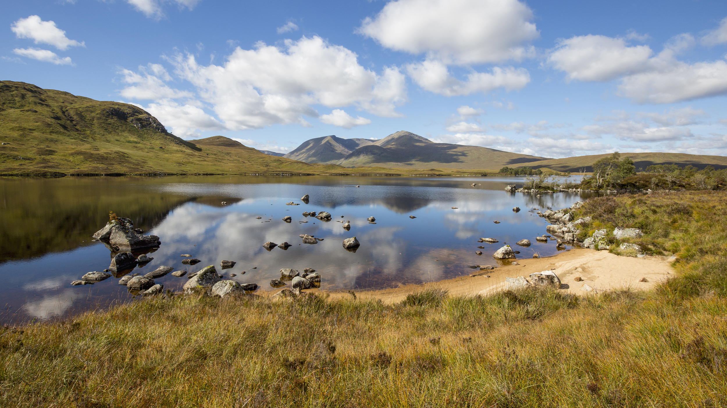A serene landscape featuring a calm lake reflecting mountains and clouds, surrounded by grassy hills and rocky shores.