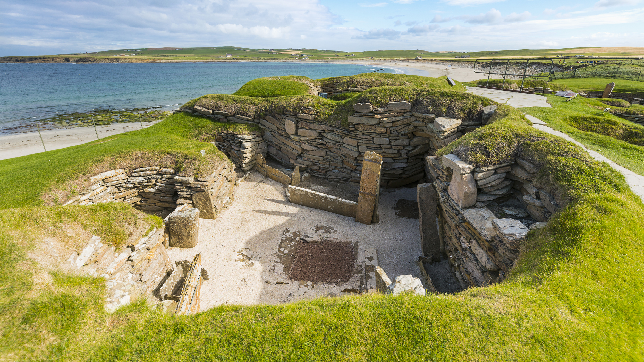 Ancient stone ruins surrounded by grassy mounds, overlooking a beach and ocean under a partly cloudy sky.