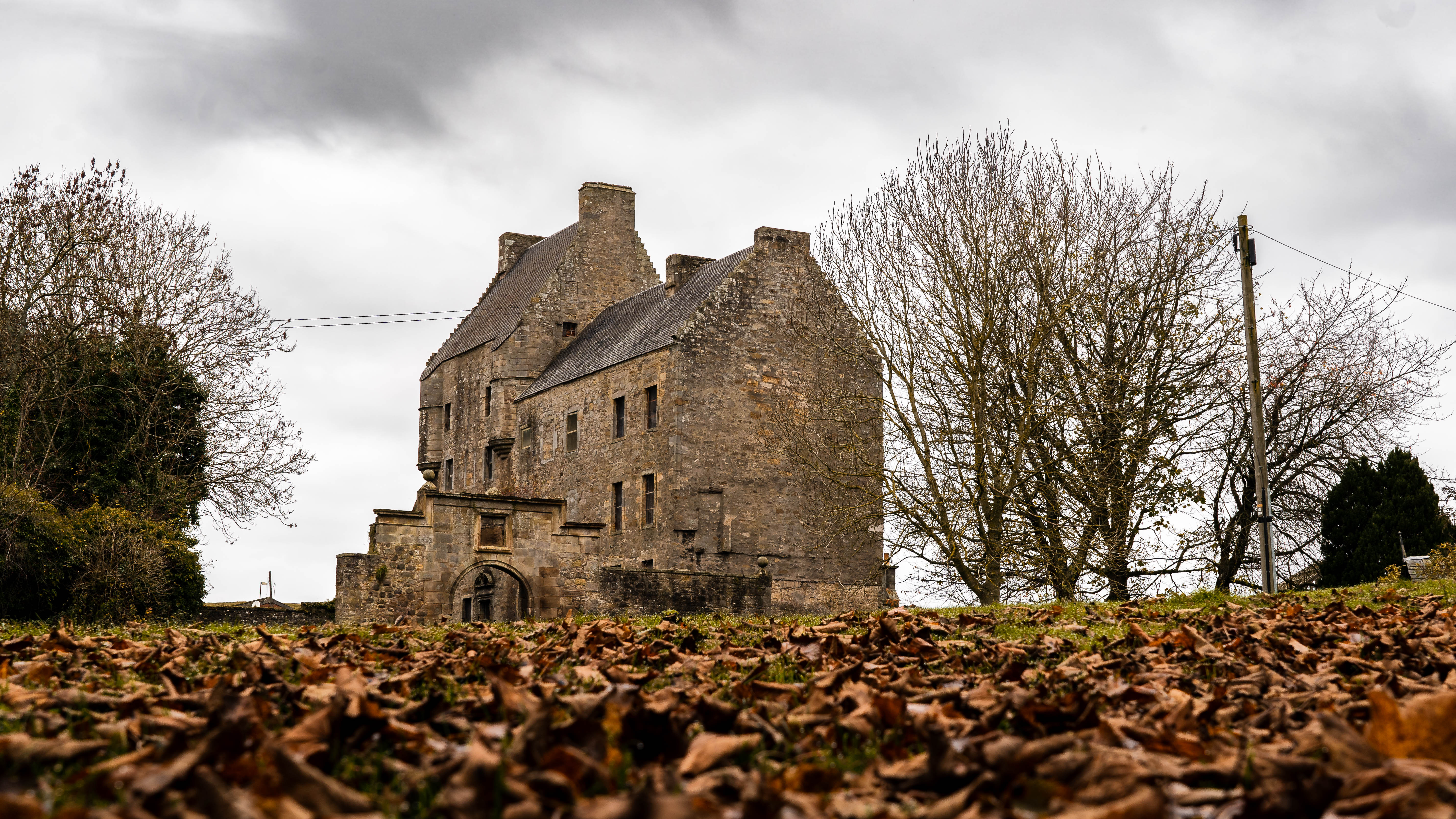 A historic stone building set against a cloudy sky, surrounded by leaf-covered ground and bare trees in the foreground.