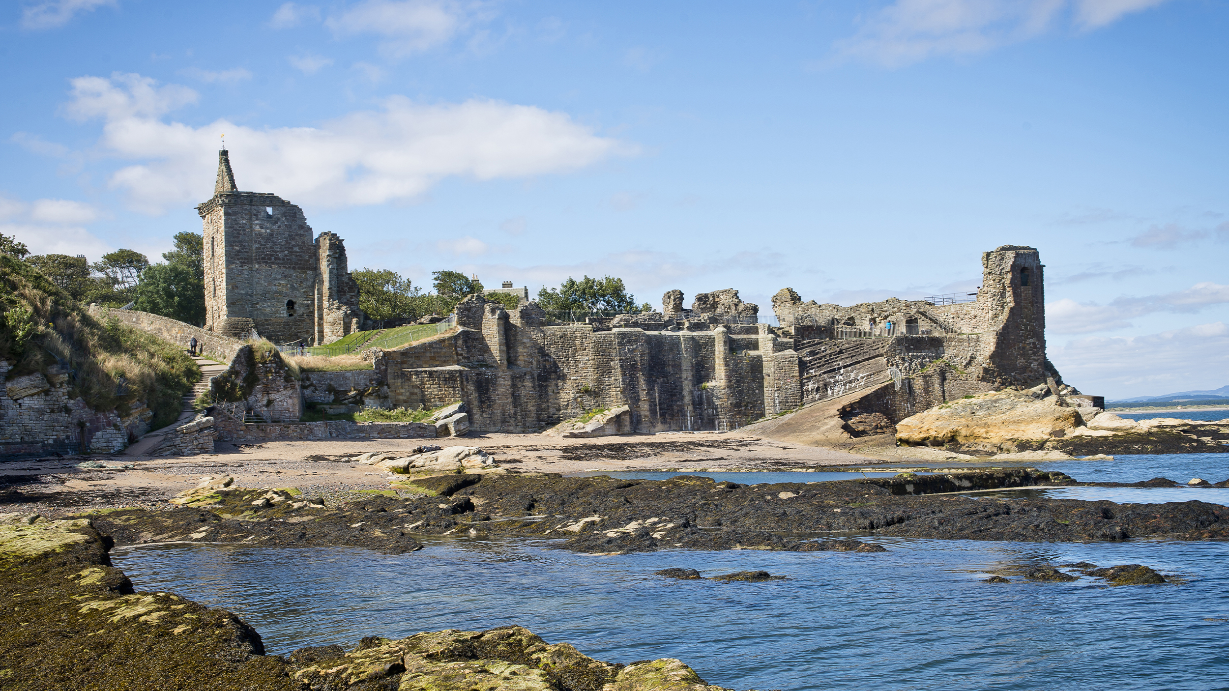 Ruins of a historic stone tower and walls by the coast, surrounded by sandy beach and rocky shoreline under a bright blue sky.