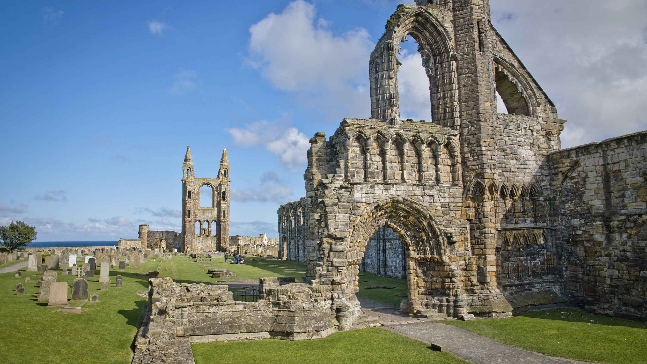 Ruins of an ancient stone abbey with towers and gravestones, set against a clear blue sky by the sea.