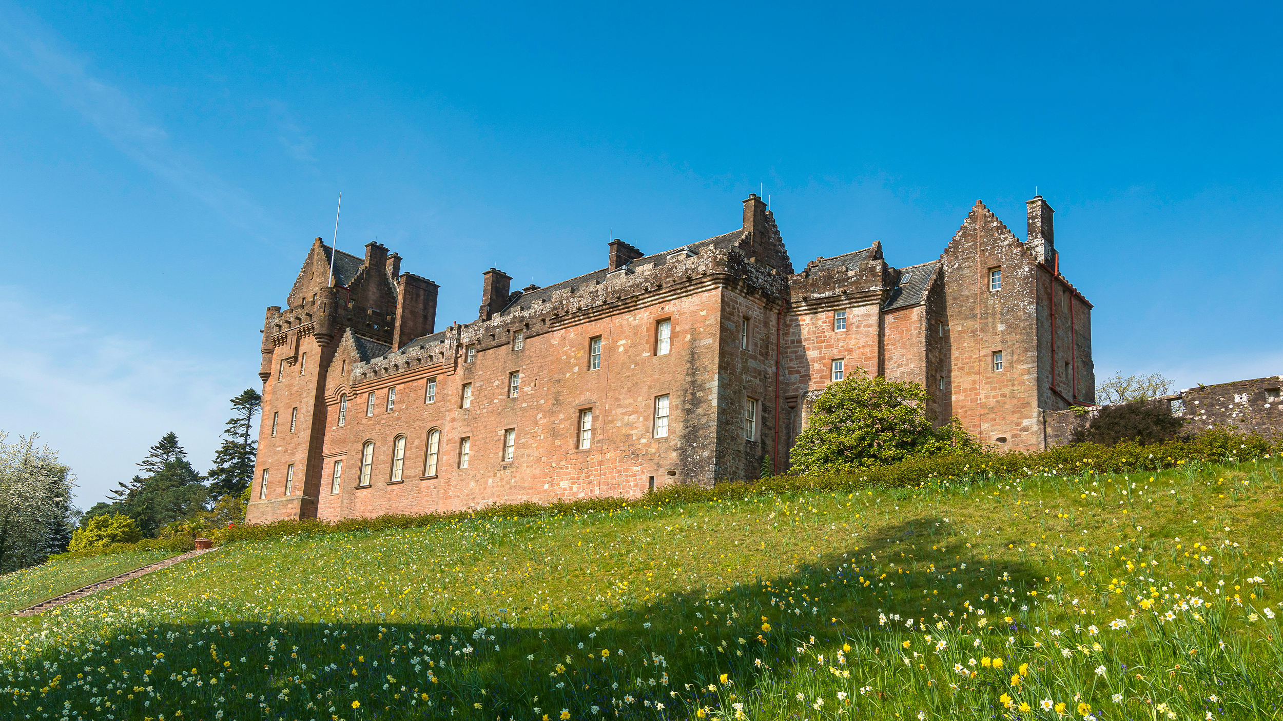 A historic castle with red stone walls surrounded by a lush green lawn and blooming flowers under a clear blue sky.