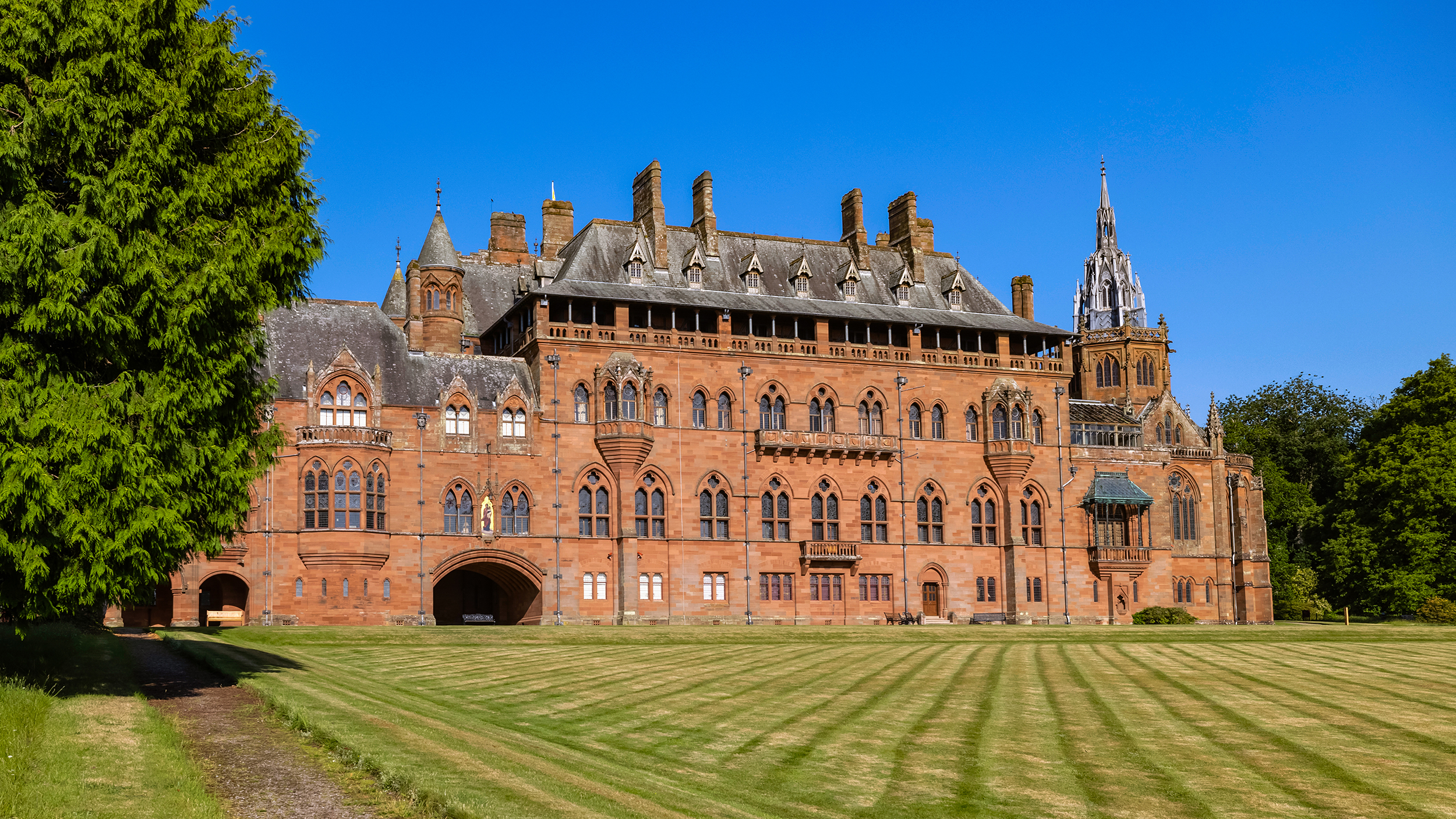 Historic red brick mansion surrounded by lush greenery and a clear blue sky, showcasing intricate architectural details.