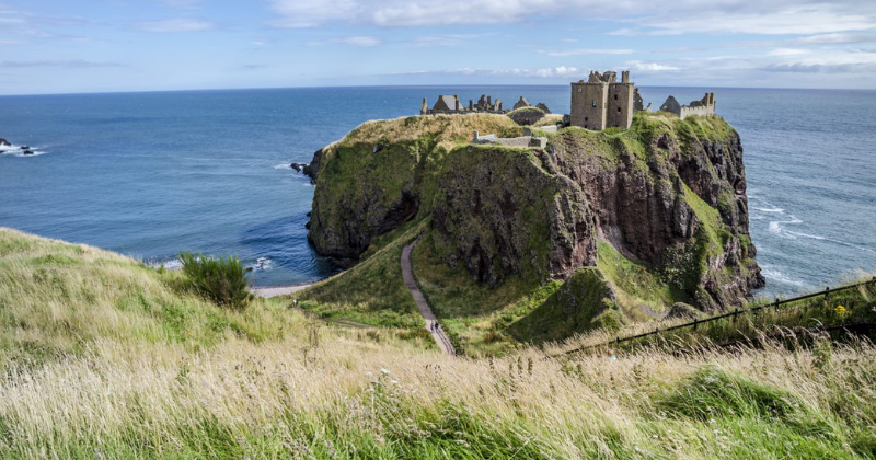A scenic view of a coastal cliff with a historic castle ruins, lush greenery, and a tranquil ocean backdrop.