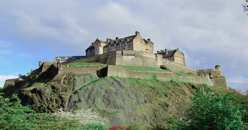 Edinburgh Castle perched atop a rocky hill, surrounded by lush greenery and dramatic skies. A historic landmark in the UK.