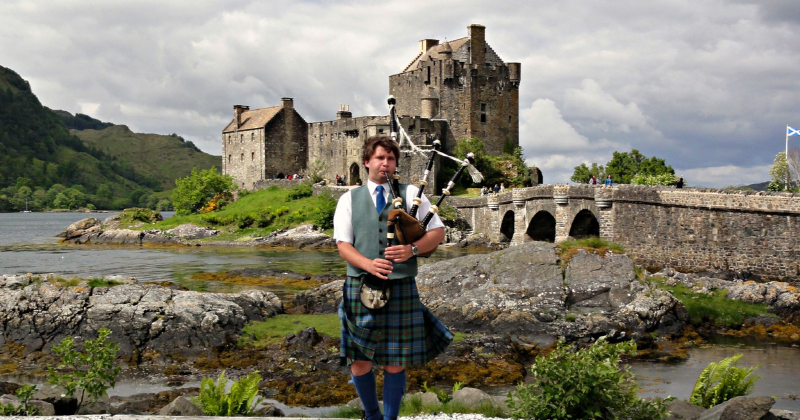 A man in a kilt plays bagpipes near a historic castle by the water, surrounded by lush greenery and dramatic skies.
