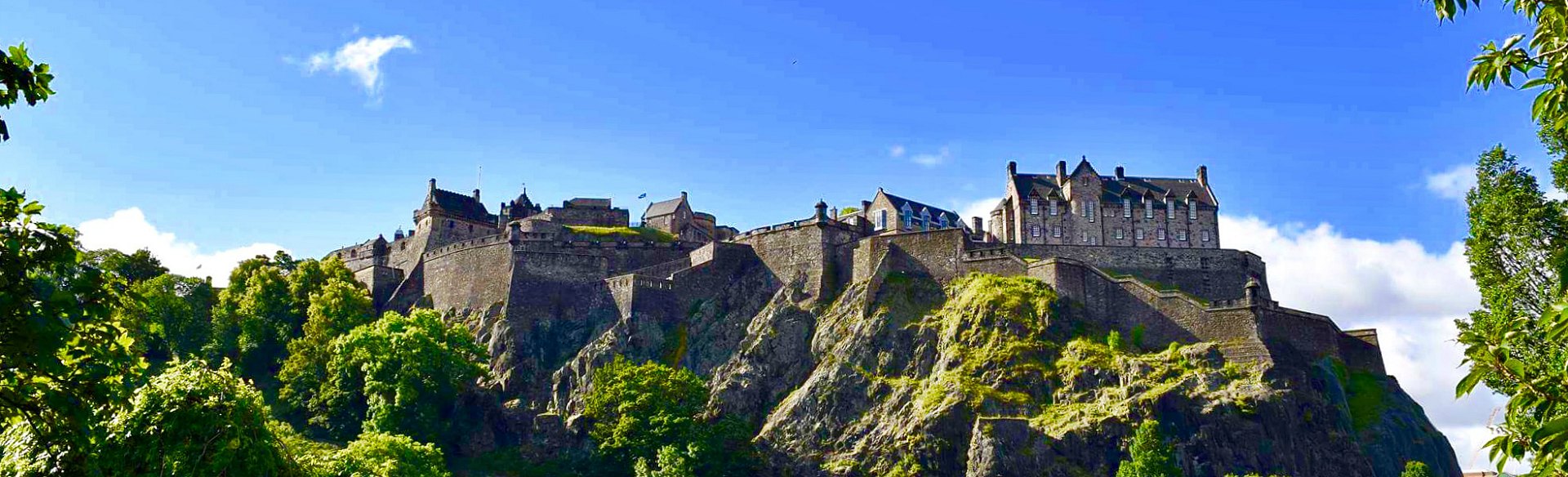 A historic castle perched on a rocky hill, surrounded by lush greenery under a bright blue sky.
