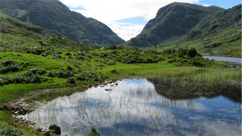 mountains surrounding a green field with a puddle showing the reflection of the blue sky