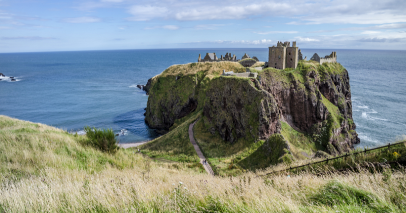 A scenic view of a castle perched on a rugged cliff overlooking the sea, surrounded by lush greenery and a clear blue sky.