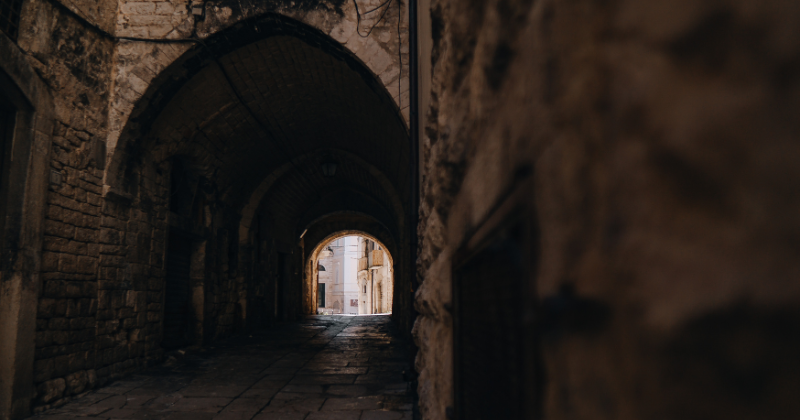 A narrow, historic alleyway with stone walls, leading to an arched entrance that opens to light. Perfect for exploring.