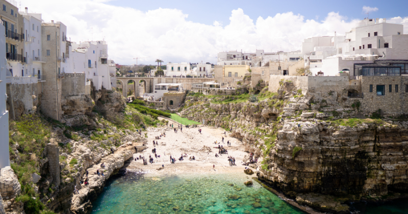 A scenic view of a rocky coastline with beachgoers, clear turquoise water, and whitewashed buildings in the background.