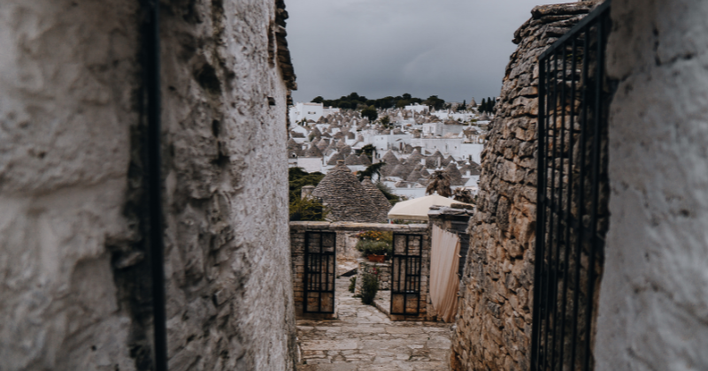Quaint stone alleyway leading to a view of whitewashed buildings and trulli in a charming village under a cloudy sky.