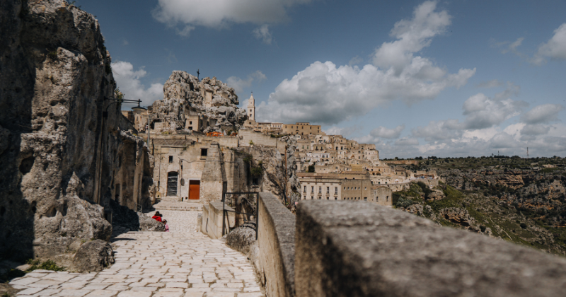 Scenic view of ancient stone buildings on a hillside, surrounded by rocky cliffs and a blue sky with scattered clouds.
