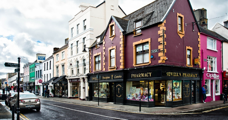 Colorful shops line a quaint street, featuring a pharmacy and various boutiques under a cloudy sky.