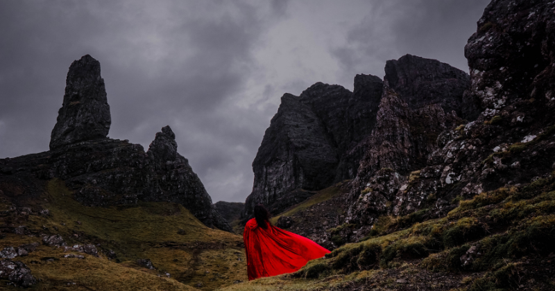A woman in a red cape standing beneath enormous dark rock formations