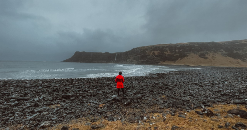 A woman in a red coat standing on a foggy, dark, rocky beach