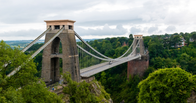 Clifton Suspension Bridge spans a lush valley, connecting two cliffs with dramatic towers and cable support under a cloudy sky.