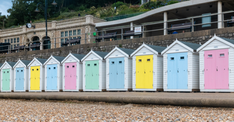 Row of colorful beach huts lined up along a pebbled shore, with a historic building in the background.