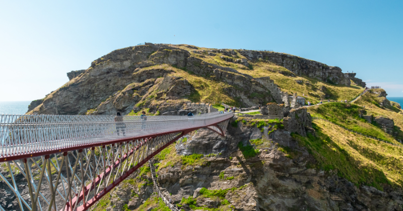 A scenic view of a bridge connecting cliffs, leading to lush green hills and rocky shores under a bright blue sky.
