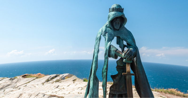 Statue of a cloaked figure holding a sword, overlooking a scenic coastline with a blue sky and ocean in the background.
