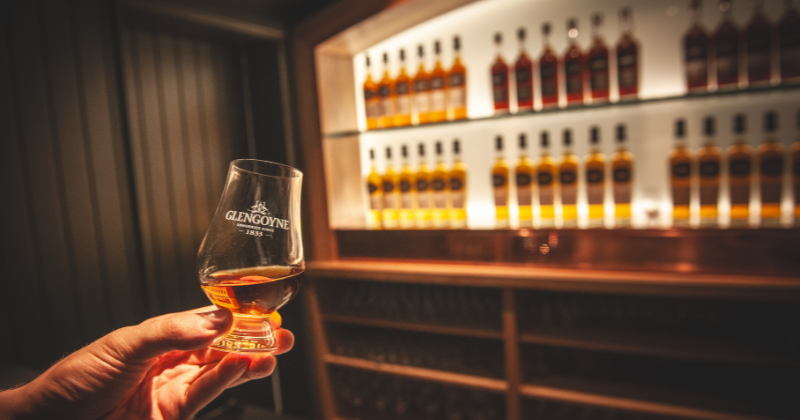 a hand holding a glass of whisky with bottles in the background