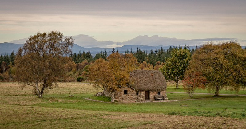 A quaint, thatched-roof cottage surrounded by autumn trees and rolling hills under a cloudy sky. Perfect for a scenic tour.
