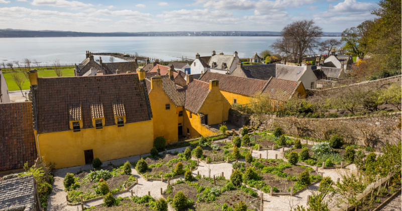 A view of yellow houses and a garden overlooking a calm sea under a partly cloudy sky. Ideal for Rabbie's Tours.