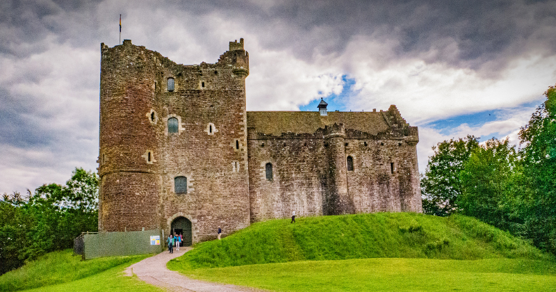 Historic stone castle on a grassy hill, surrounded by trees, under a cloudy sky with visitors exploring the site.