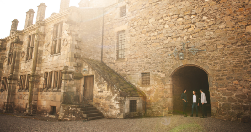 Visitors explore the historic stone exterior of a castle, with a sunny ambiance and a staircase leading to an archway.