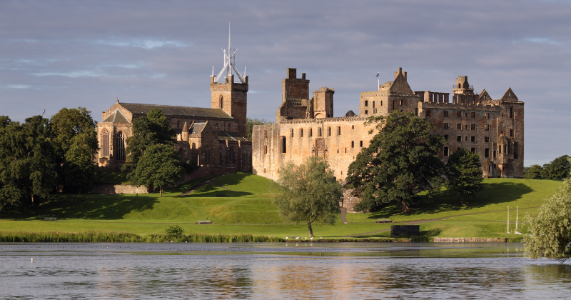 A historic castle sits atop a green hill alongside a calm lake, surrounded by trees and under a partly cloudy sky.