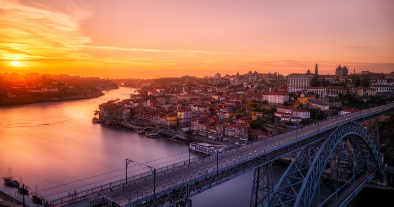 Steel bridge in front with a river running through it and the orange hued sky in the background as the sun sets
