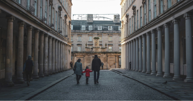 A family walks hand-in-hand down a historic cobbled street lined with columns, enjoying a peaceful day.