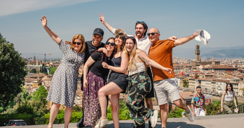A joyful group of eight people poses together with arms raised, smiling against a scenic city backdrop.