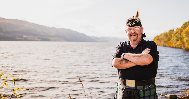 driver-guide with a feather in hat crossed arms standing on the banks of a loch