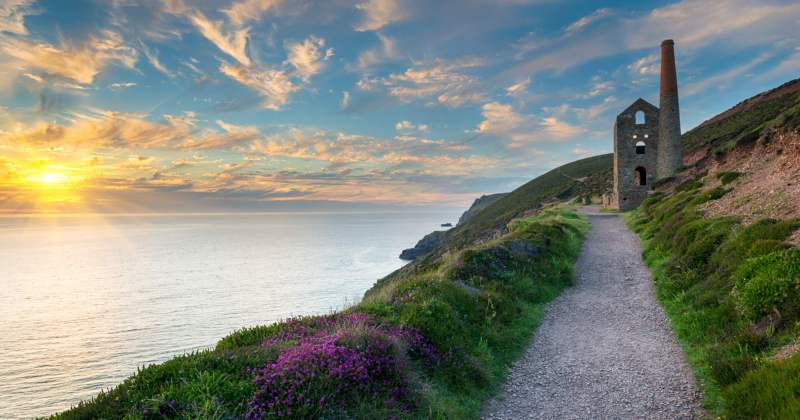 coastal path with the sun setting over the sea and a small stone building at the end of the path