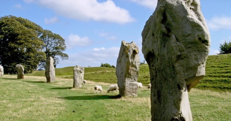 Ancient standing stones in a lush green landscape, with sheep grazing under a clear blue sky.