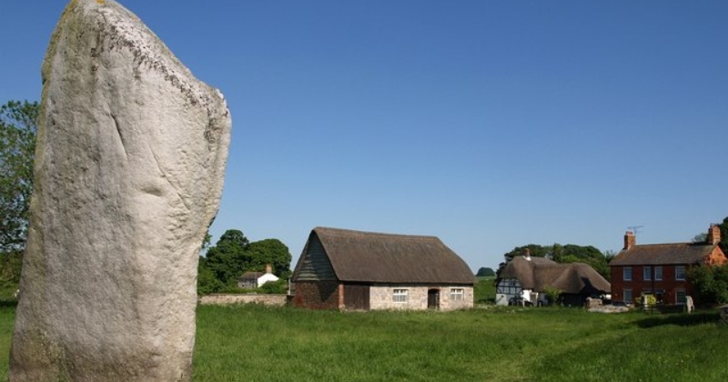 Historic stone stands in a green field with traditional thatched-roof cottages and a clear blue sky in the background.