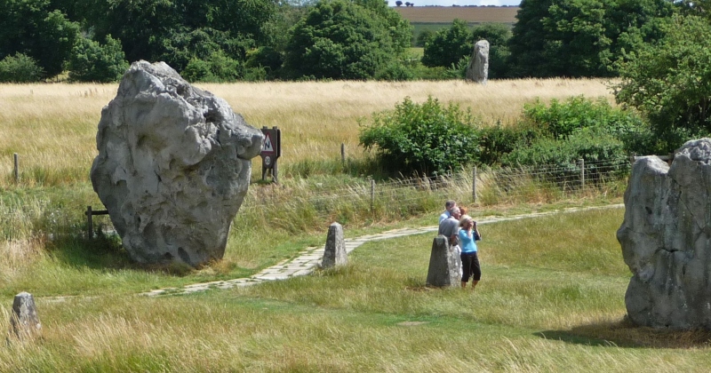 Visitors explore a prehistoric stone circle in a grassy landscape, surrounded by tall trees and open fields.