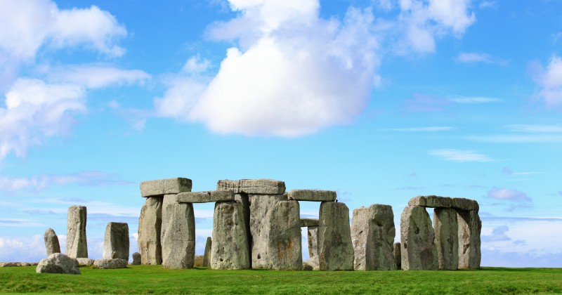 Stonehenge stands majestically on green grass under a bright blue sky dotted with clouds, showcasing its iconic prehistoric stones.