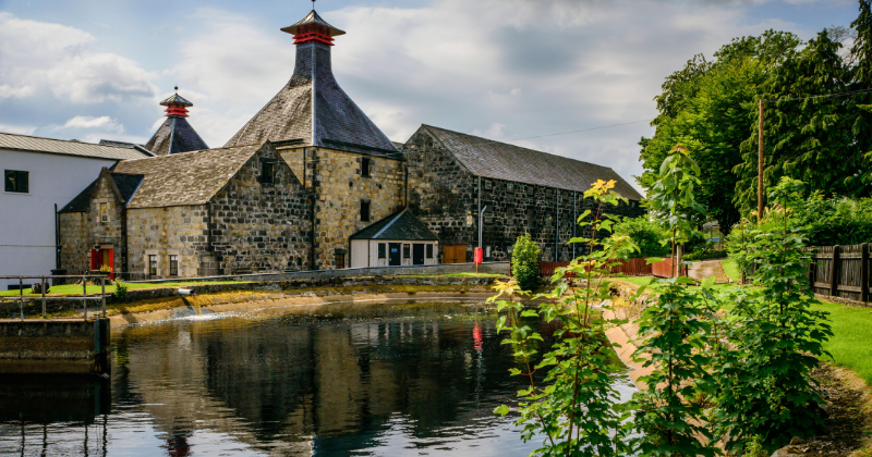 A historic stone building with red-topped towers beside a calm waterway, surrounded by lush greenery and a clear sky.