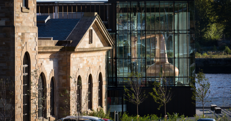 A blend of historic stone architecture and modern glass featuring a large copper distillation still by a riverside.