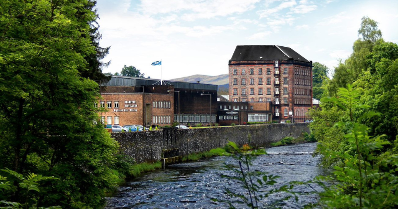 Historic brick building beside a flowing river, framed by lush greenery and the Scottish flag waving in the background.