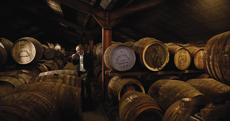 A person examines whiskey barrels in a dimly lit distillery warehouse, surrounded by stacked oak casks.